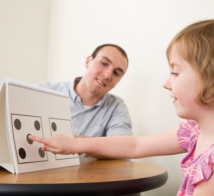 A little girl is sitting at a table with a man pointing at a board with dots on it
