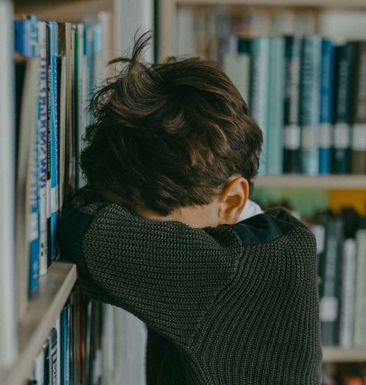 A young boy is leaning against a bookshelf with his head in his hands.