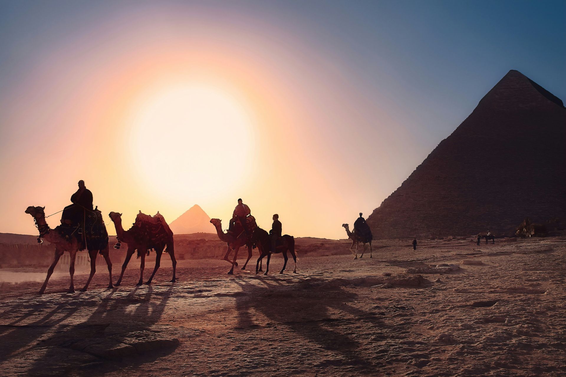 A caravan of camels walking past the massive Giza Pyramids against a bright golden sun