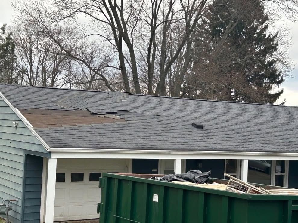 Damaged roof with missing shingles above a garage, with a green dumpster in front
