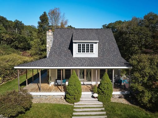 White cottage with porch and dormer window on a grassy hill, surrounded by trees under a clear blue sky