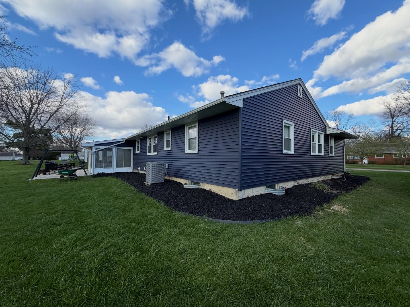 Blue house with a white porch and dark mulch, set on a green lawn under a cloudy sky