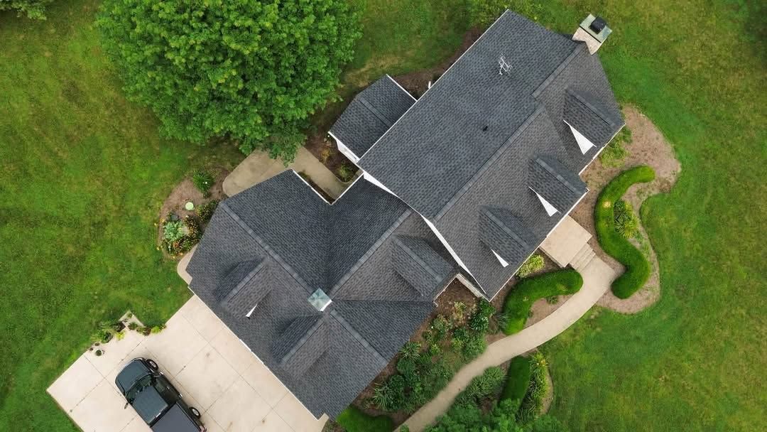 Aerial view of a gray-roofed house with a driveway, trees, and green lawn.