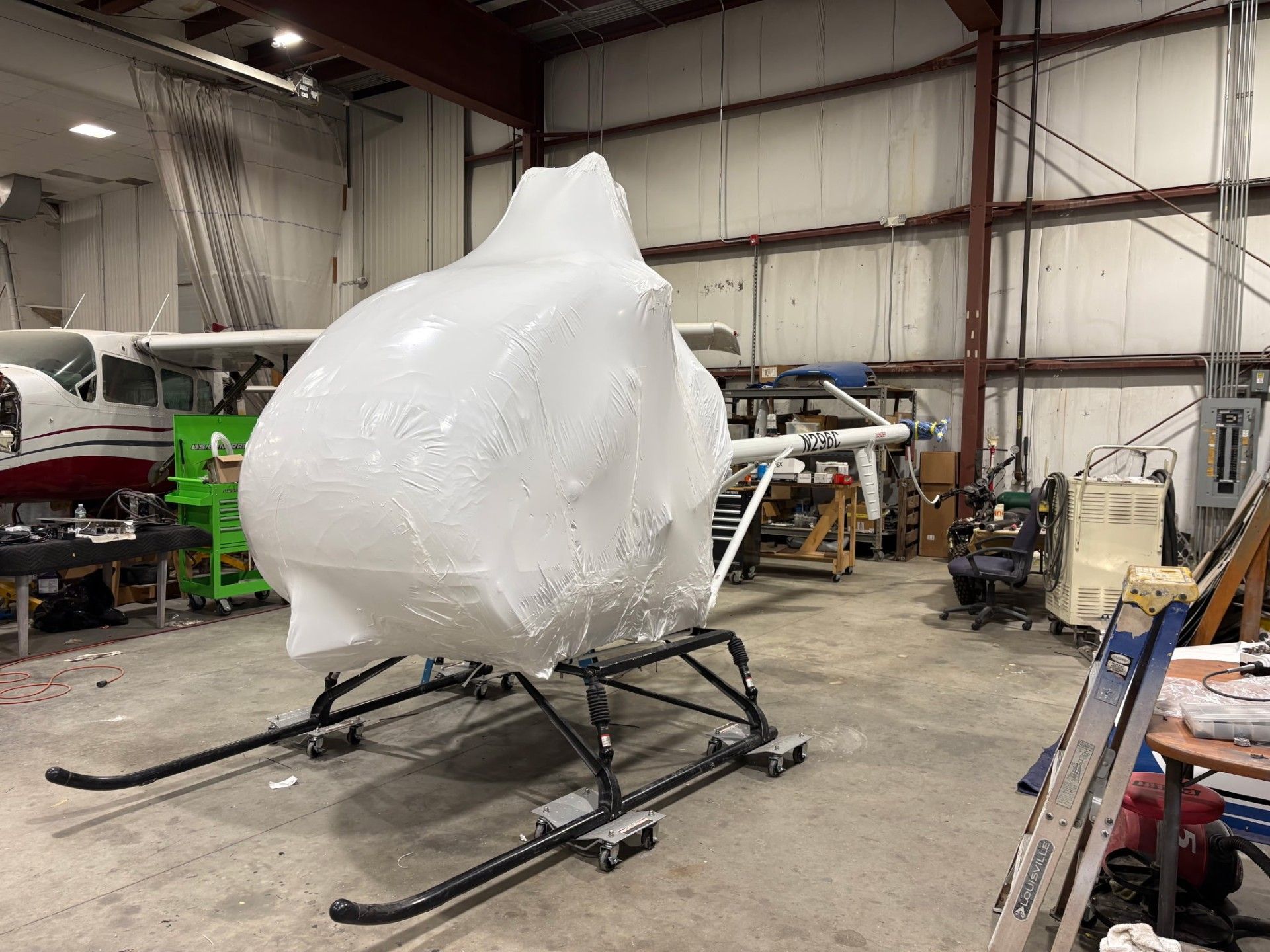 A small helicopter, wrapped in white plastic, sits in a large hangar.