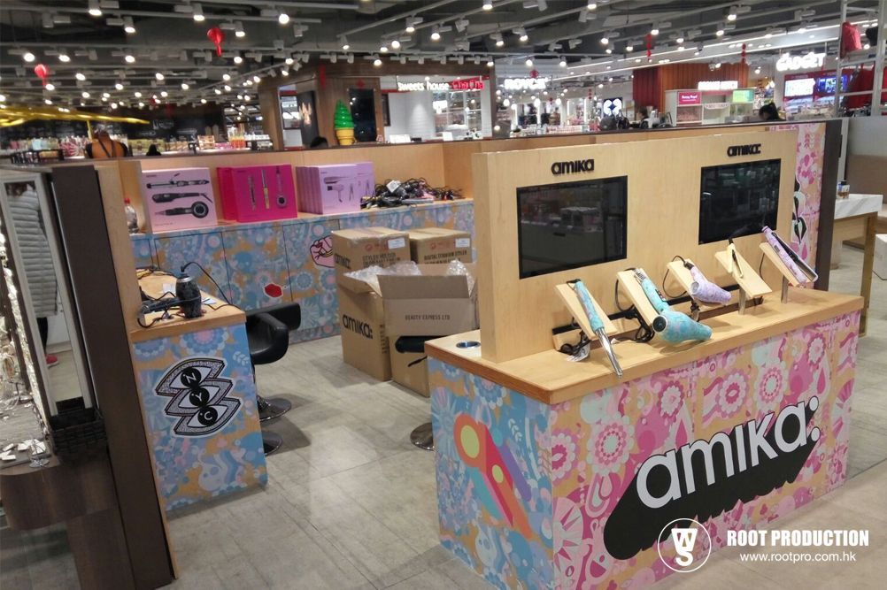 Amika product display in a store. Colorful booth with hair styling tools and branding; boxes in background.