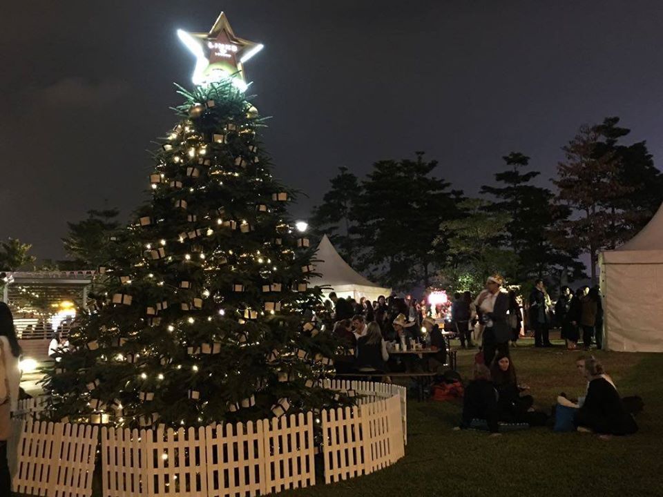Lit Christmas tree with star topper, white picket fence, crowd gathering outdoors at night.