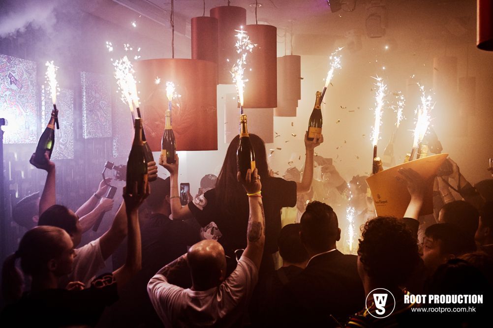 People celebrating in a club, holding champagne bottles with sparklers. Dim lighting, red/orange glow.