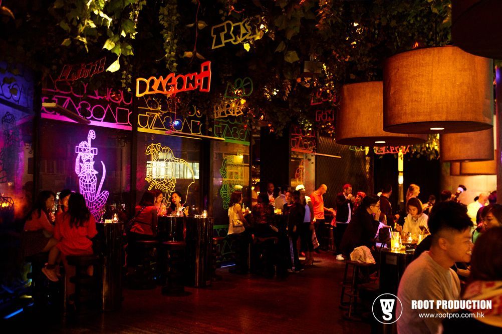 Interior of a dimly lit bar with patrons, neon signs, and hanging lamps.