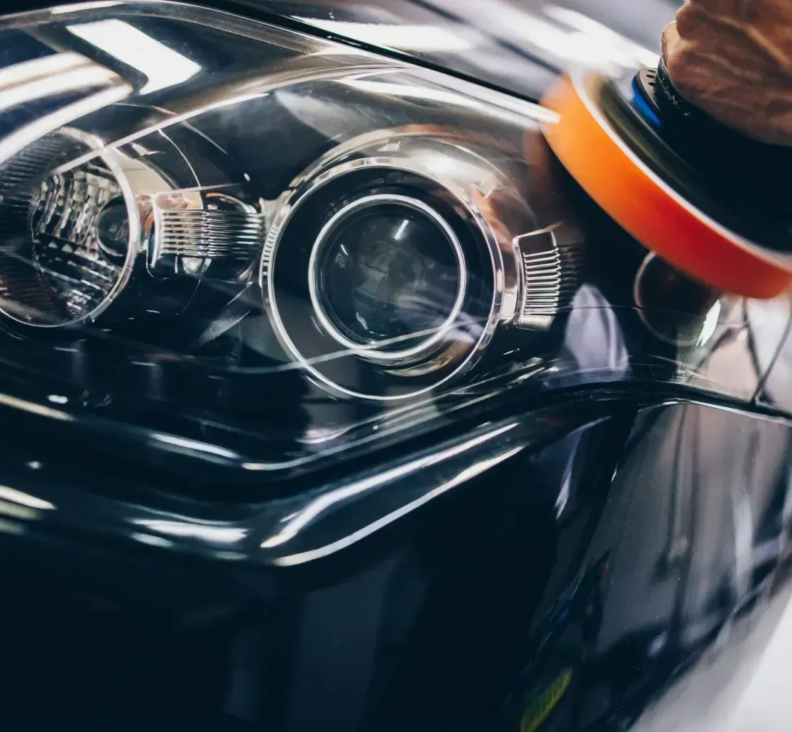 A hand uses a motorized buffer tool to polish the glossy, dark headlight and hood of a car.