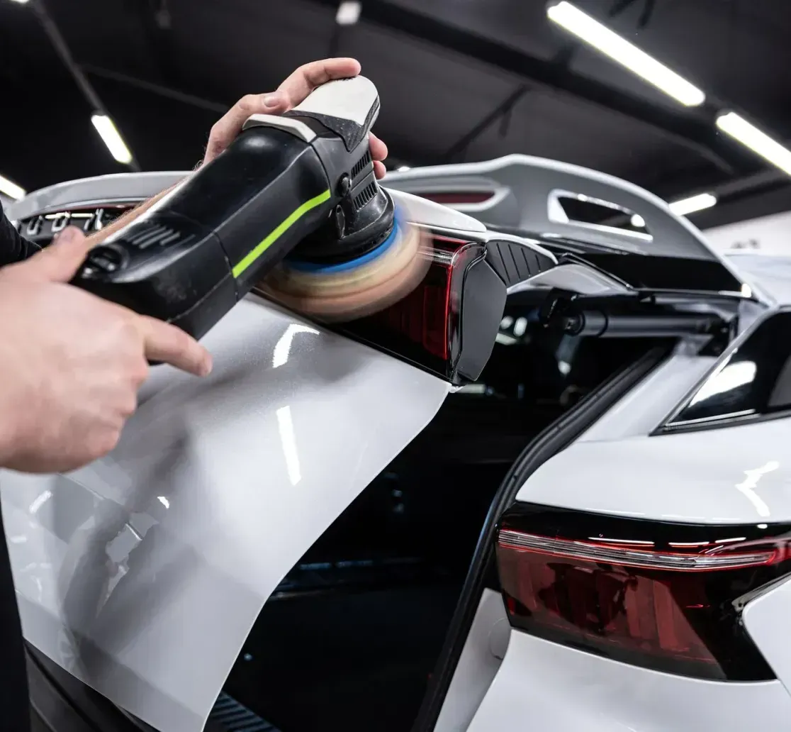 A person uses a power polisher on the white trunk of a car inside a brightly lit workshop.