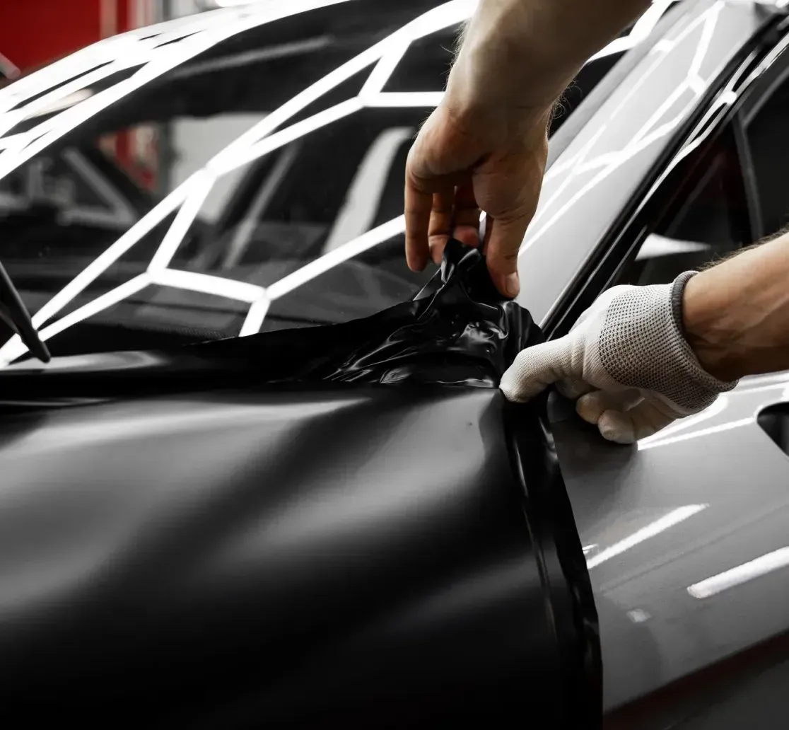 A person wearing a glove carefully applies a black matte vinyl wrap to the side panel of a car in a workshop.