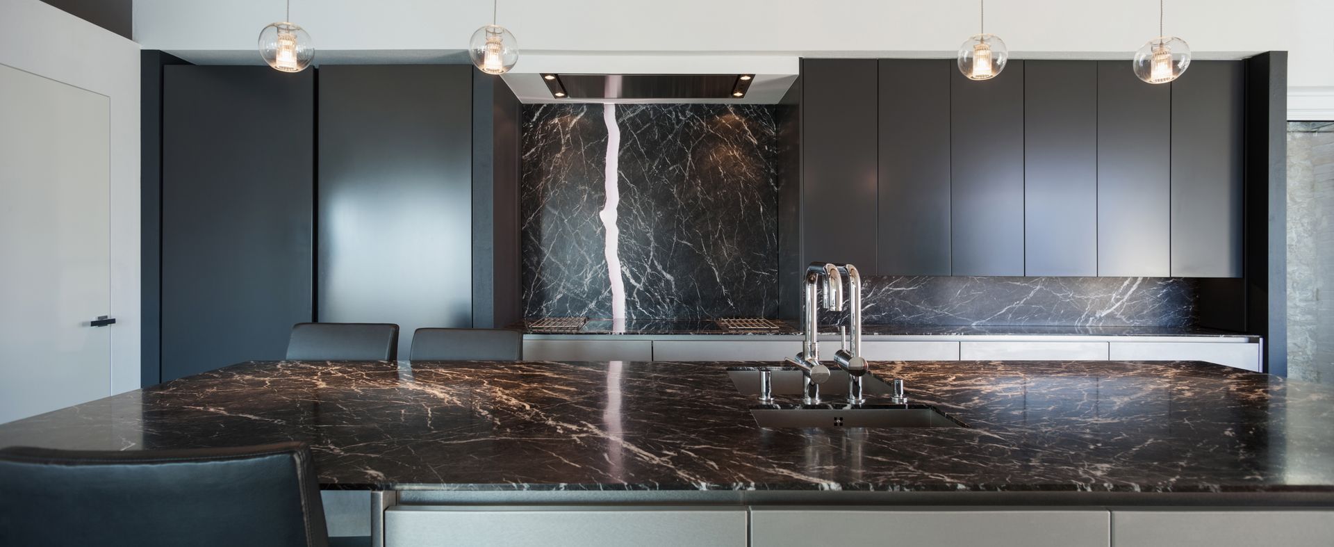 A modern kitchen featuring a dark marble island and backsplash with sleek black cabinetry and hanging pendant lights.