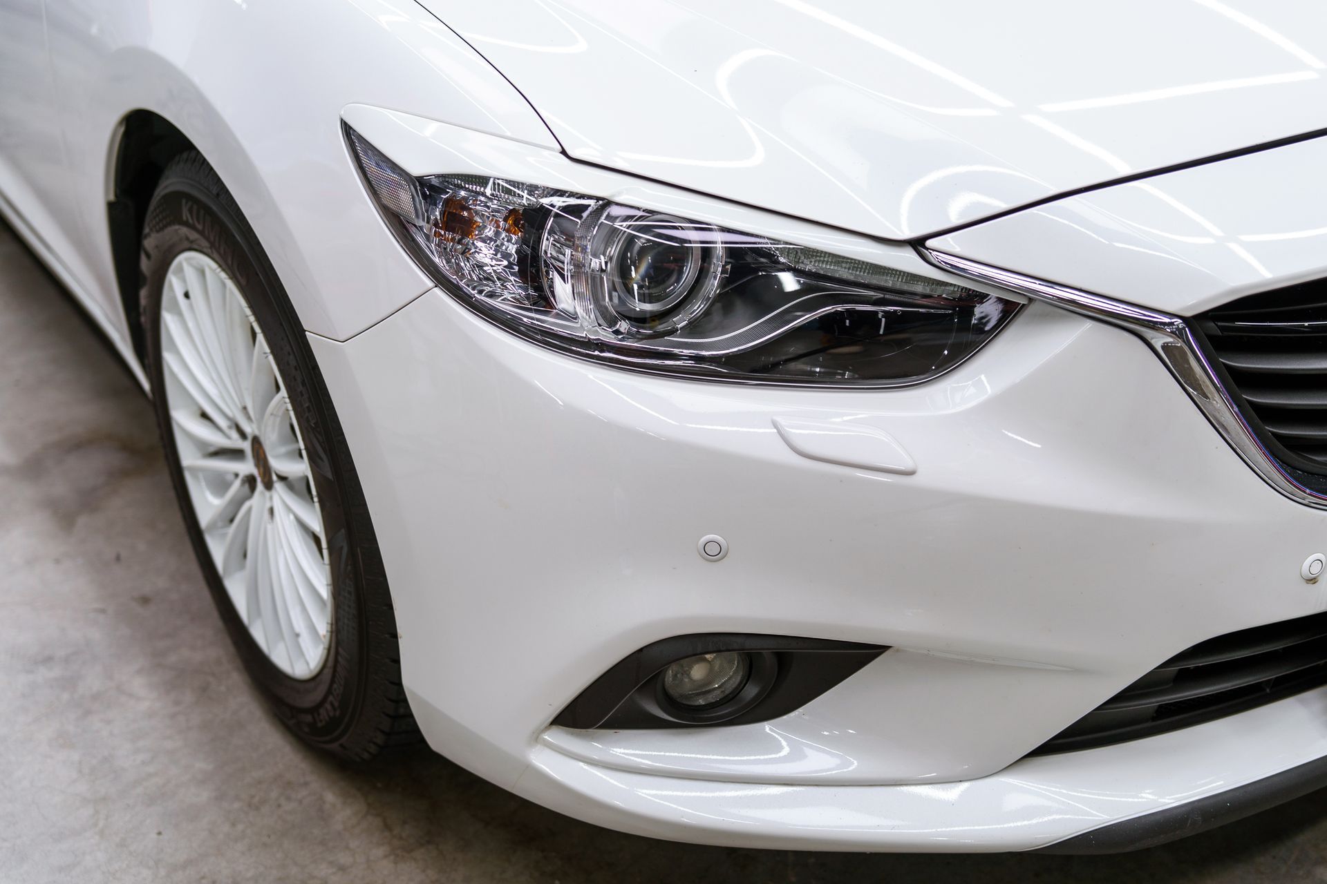 Front view of a white Mazda 6 sedan focusing on the headlight, wheel, and bumper.
