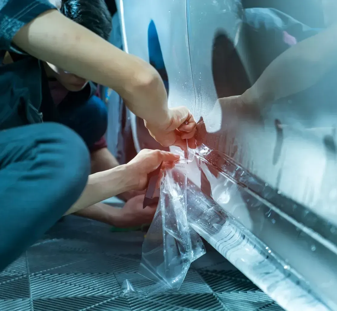 A technician carefully applying a clear protective film to the side panel of a shiny vehicle in a workshop.