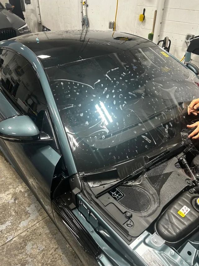 A person applies clear paint protection film to the hood of a dark gray car inside a garage.