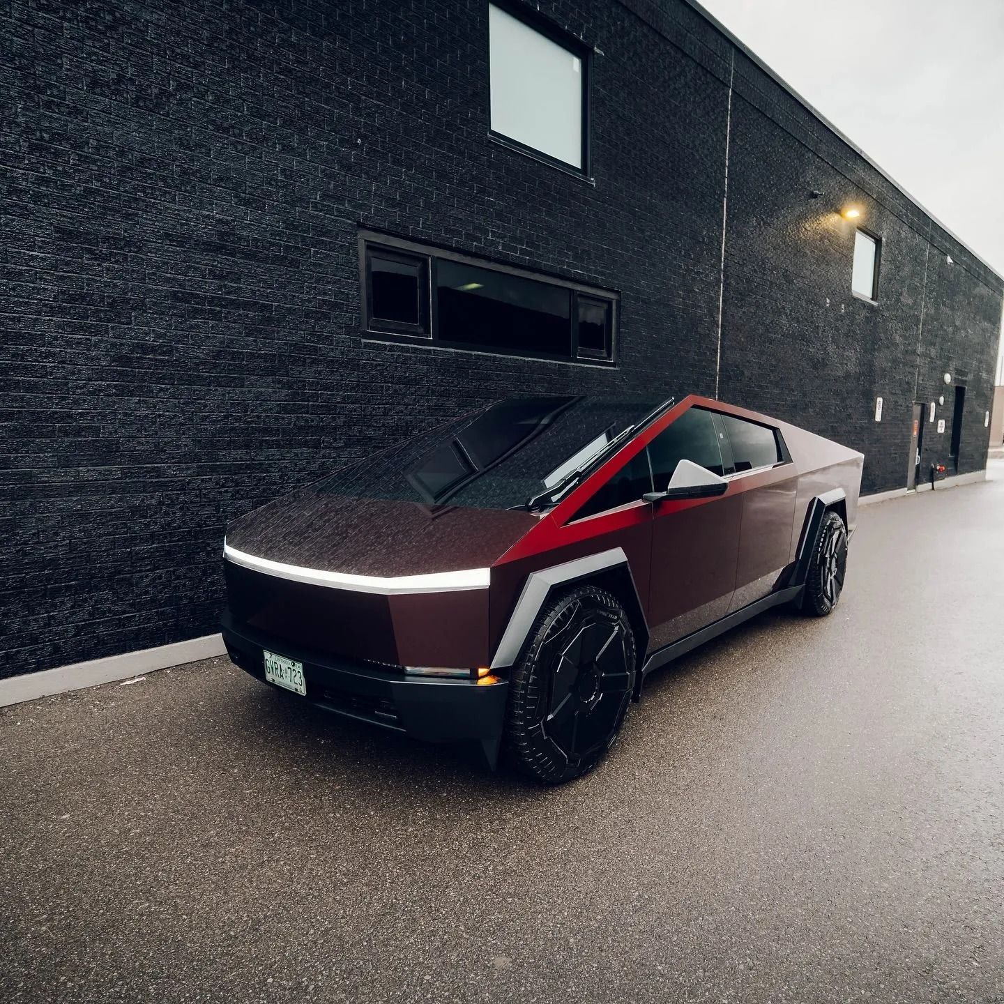 A deep red Tesla Cybertruck parked on a paved lot next to a dark brick building.