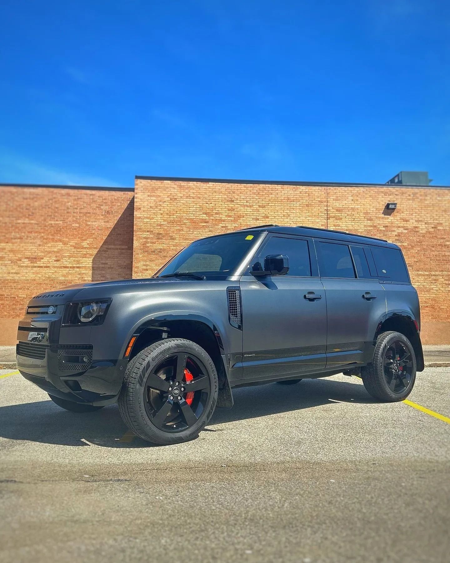 A matte dark gray Land Rover Defender parked on a gravel lot in front of a brick building under a clear blue sky.