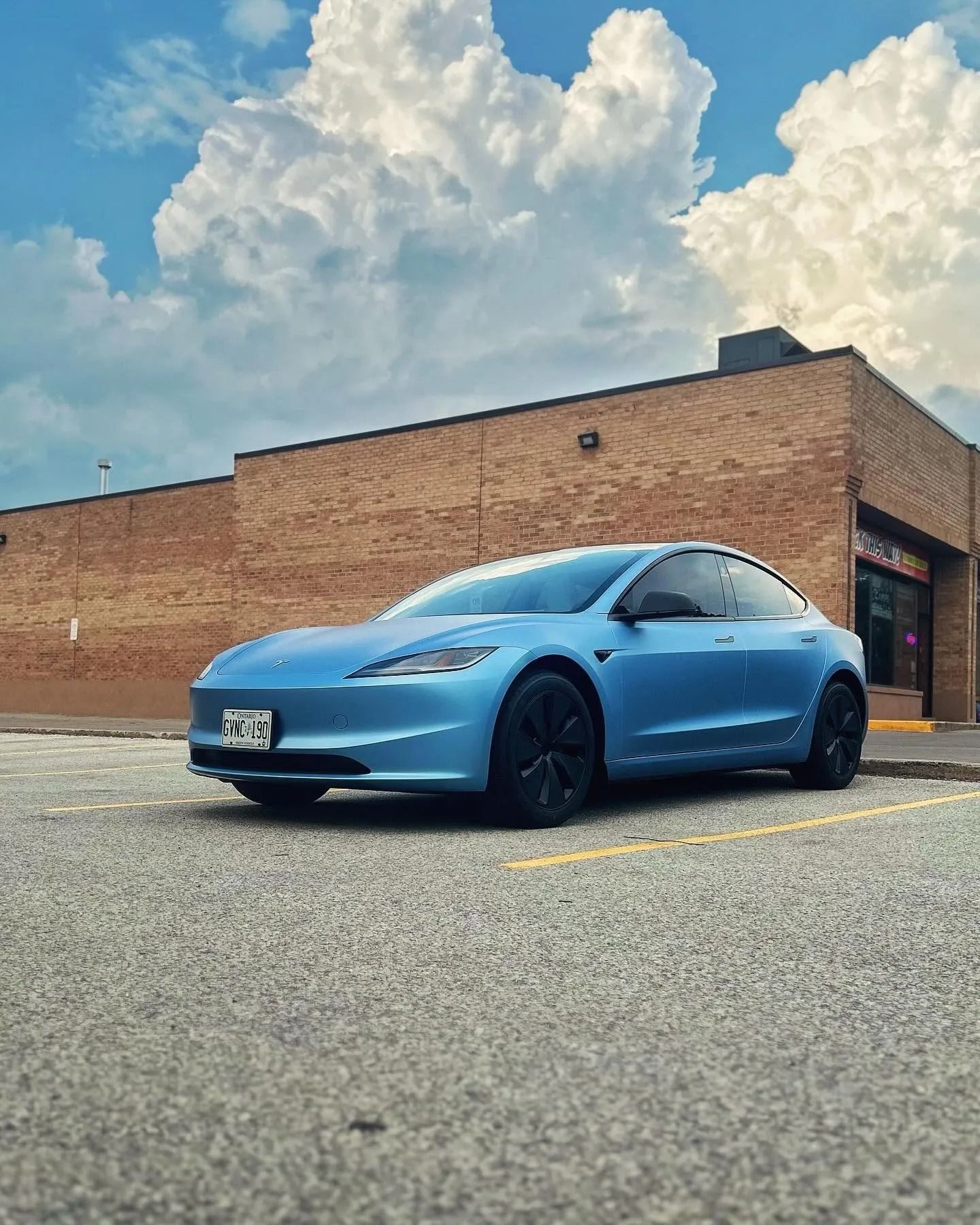 A light blue Tesla Model 3 parked in an empty asphalt parking lot in front of a tan brick building under a cloudy sky.