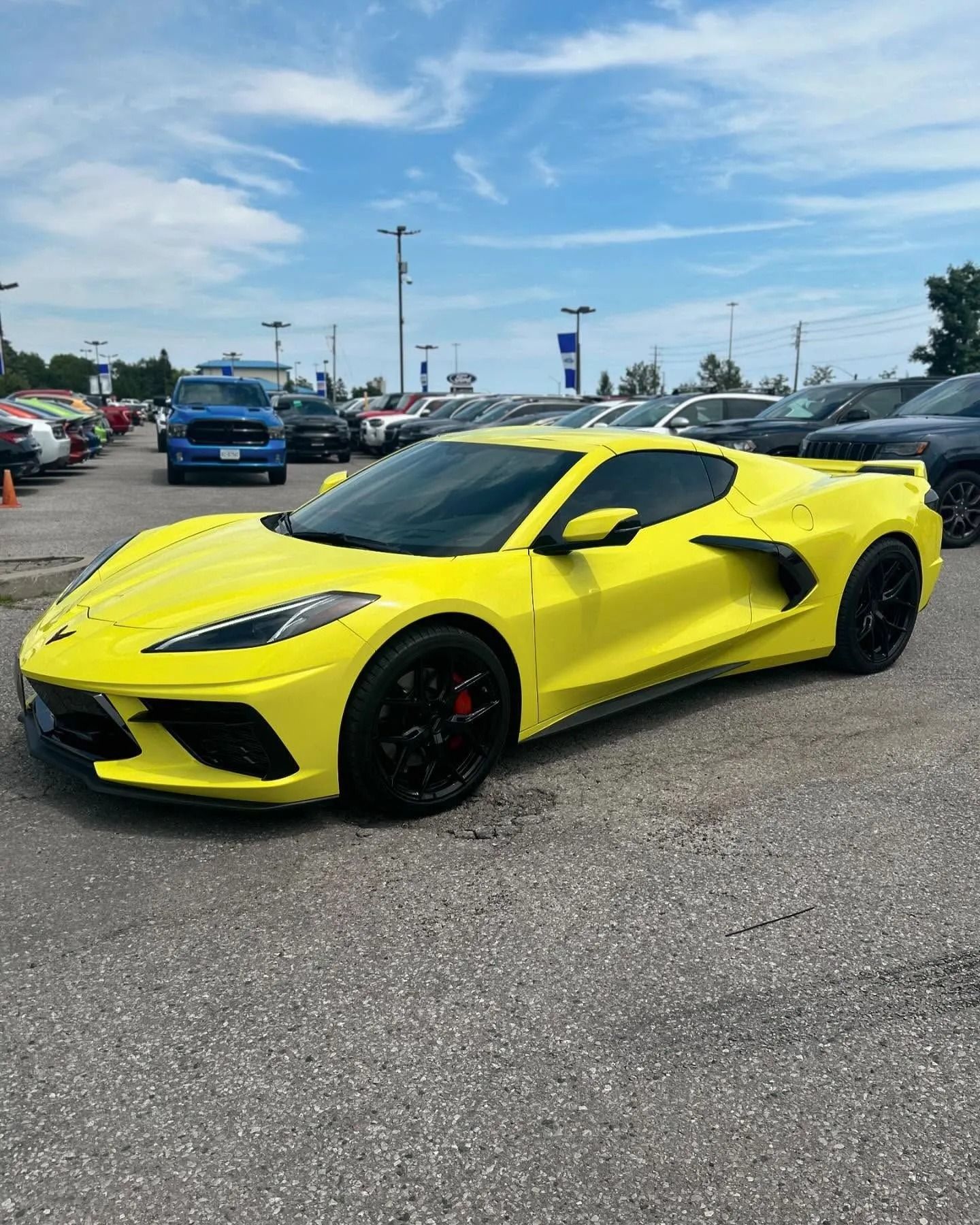 A bright yellow Chevrolet Corvette parked at an outdoor car dealership under a clear blue sky.