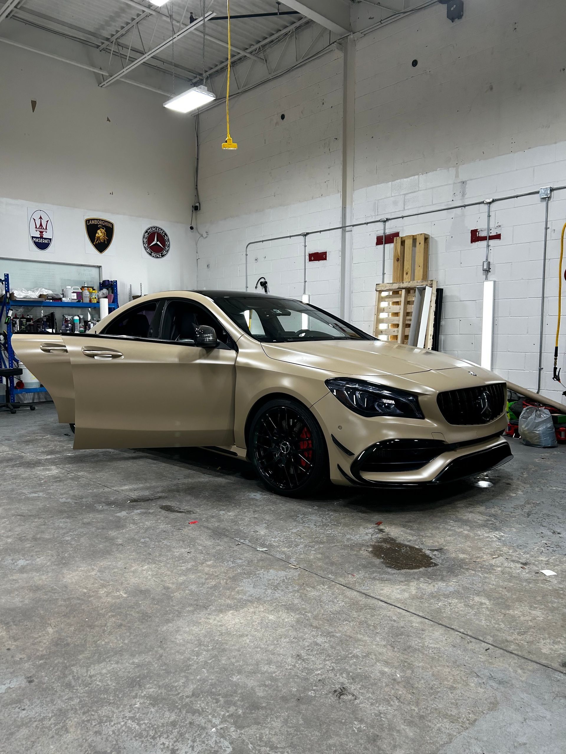 A tan Mercedes-Benz CLA with black wheels and trim, parked inside a garage with its driver-side door open.