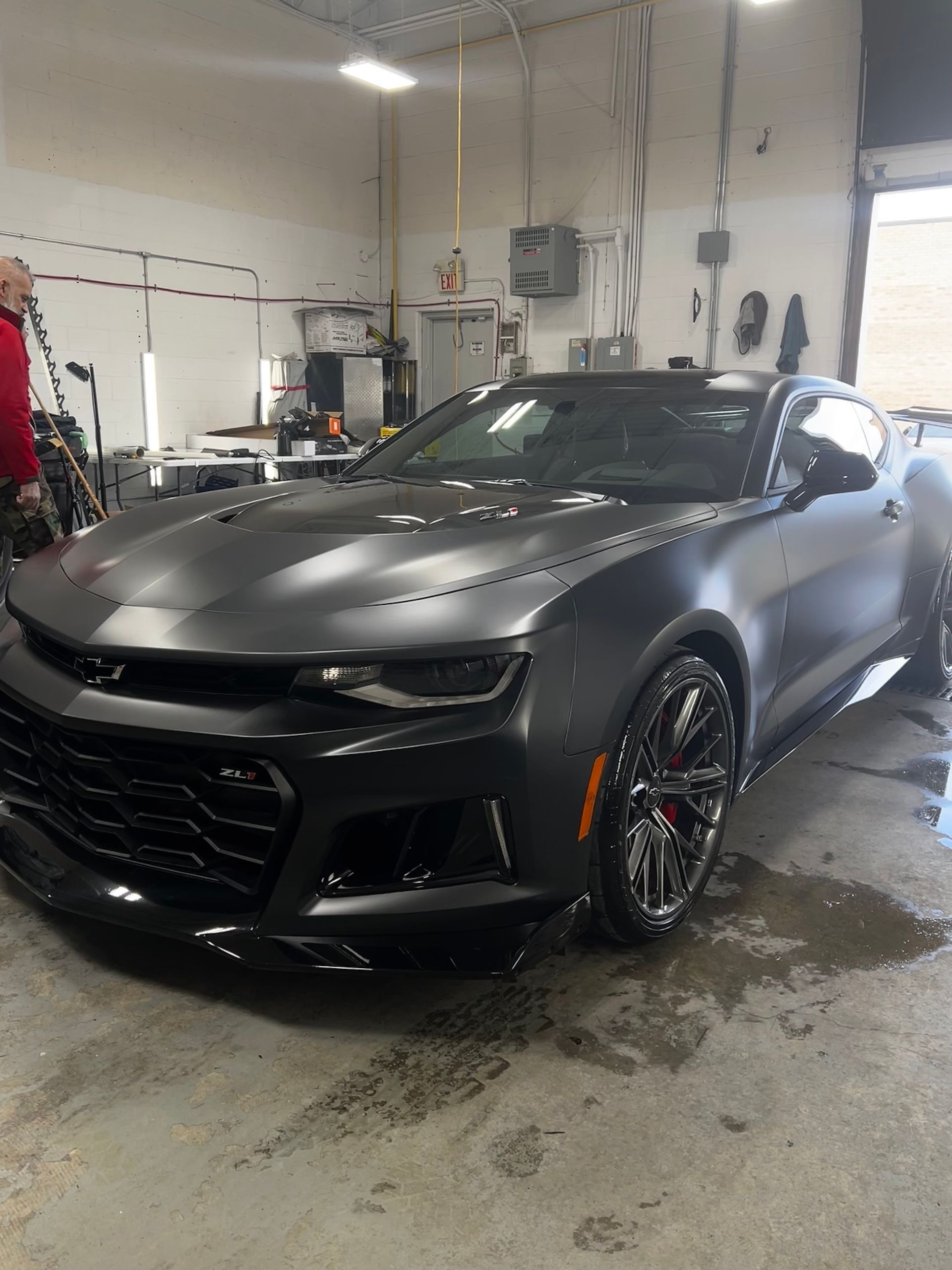 A matte black Chevrolet Camaro parked inside a garage, with a person in a red shirt visible on the left side.