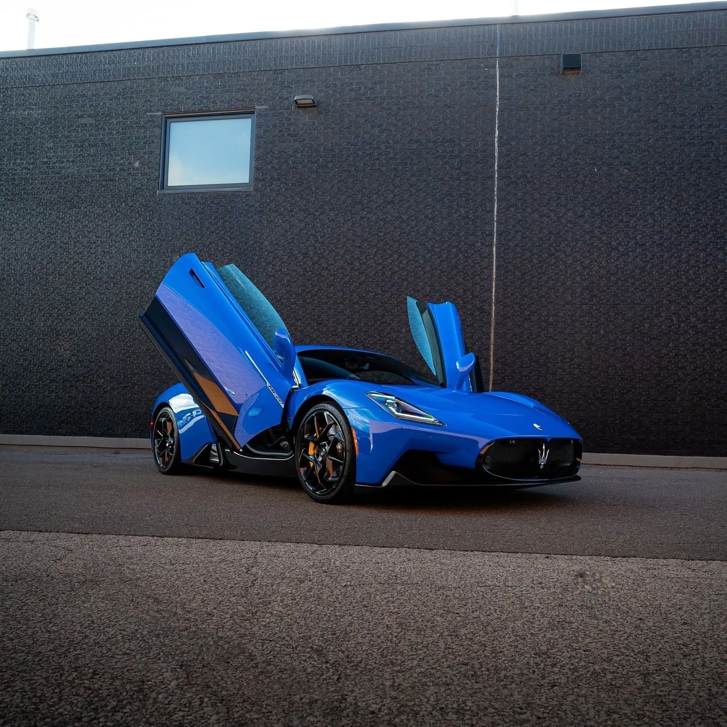 A vibrant blue Maserati MC20 sports car parked in front of a dark, textured wall with its butterfly doors wide open.