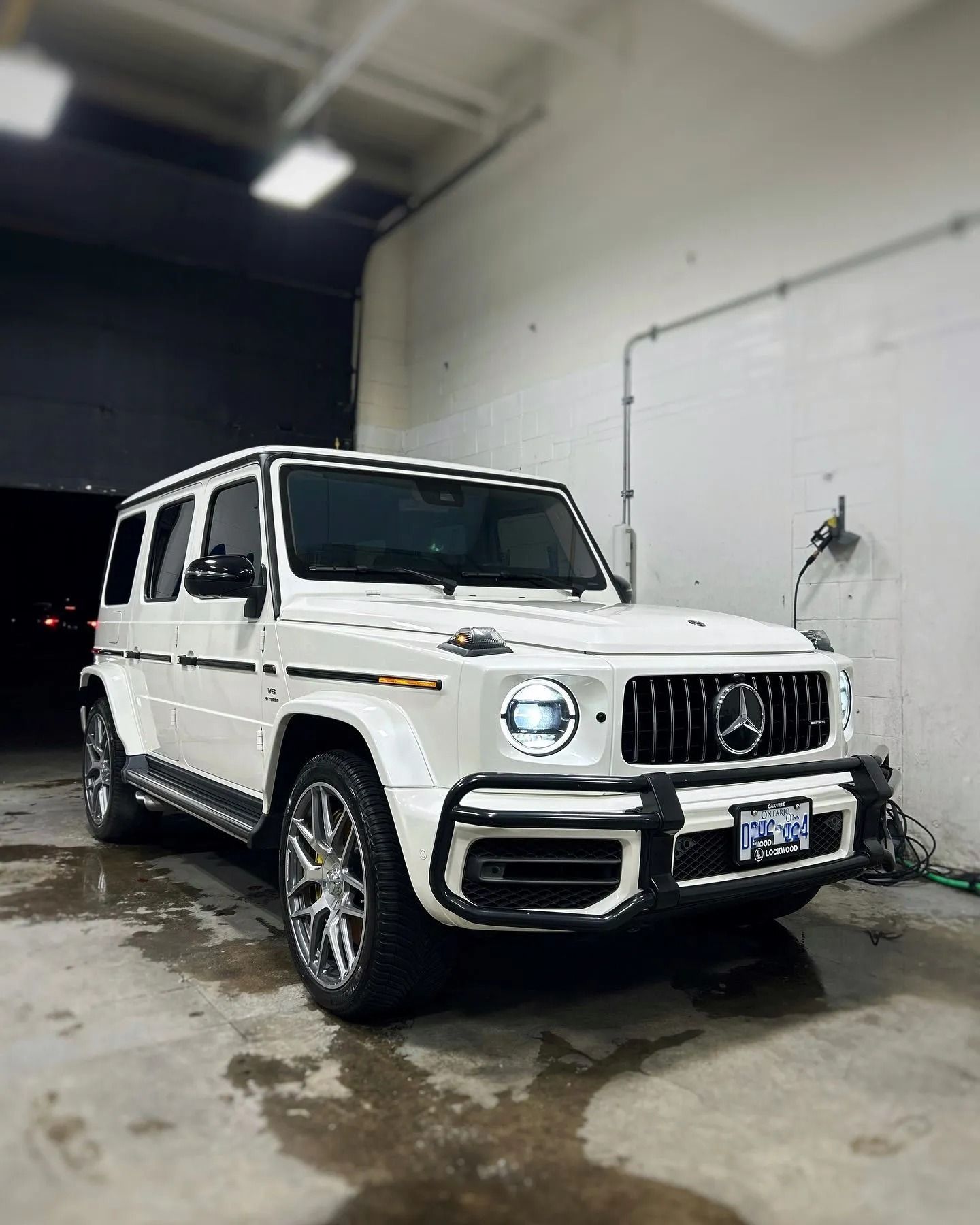 A white Mercedes-Benz G-Wagon parked inside a garage with bright lights overhead and a wet concrete floor.