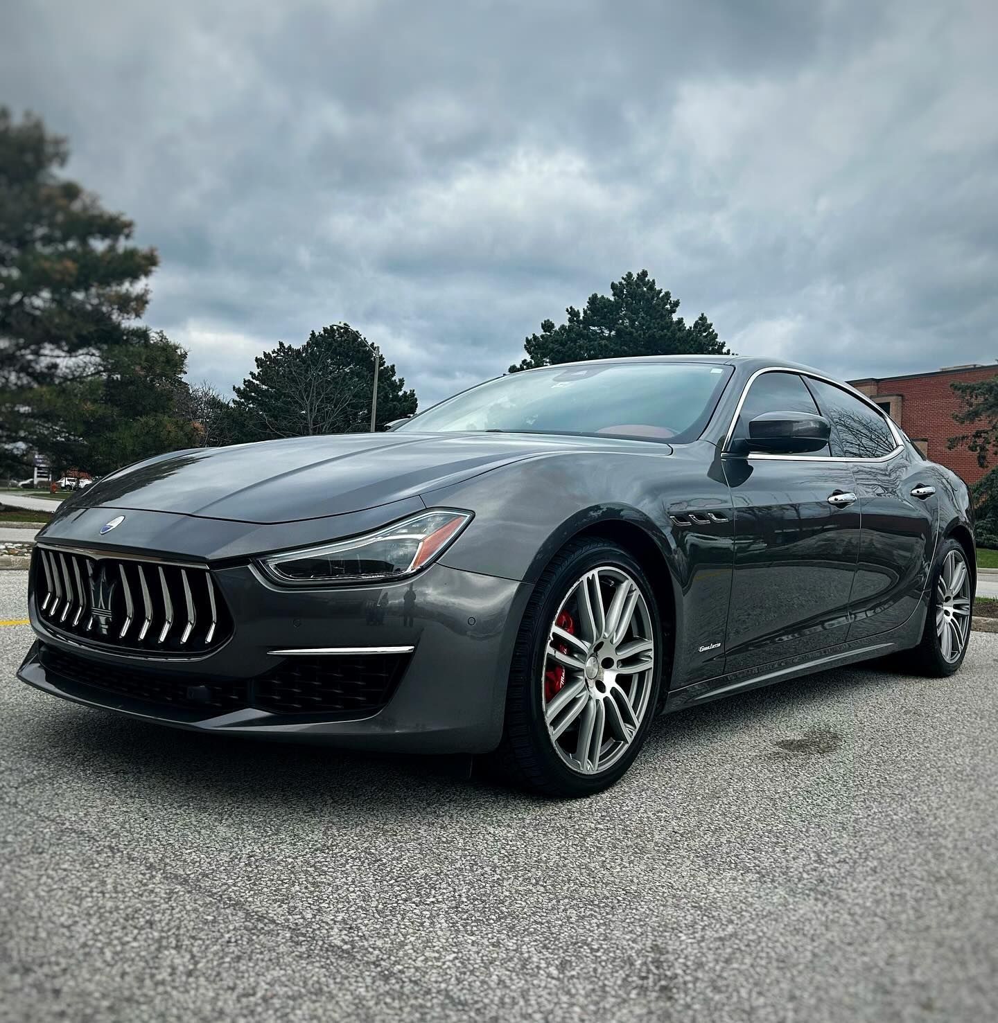 A gray Maserati Ghibli parked on an asphalt lot under a cloudy sky.