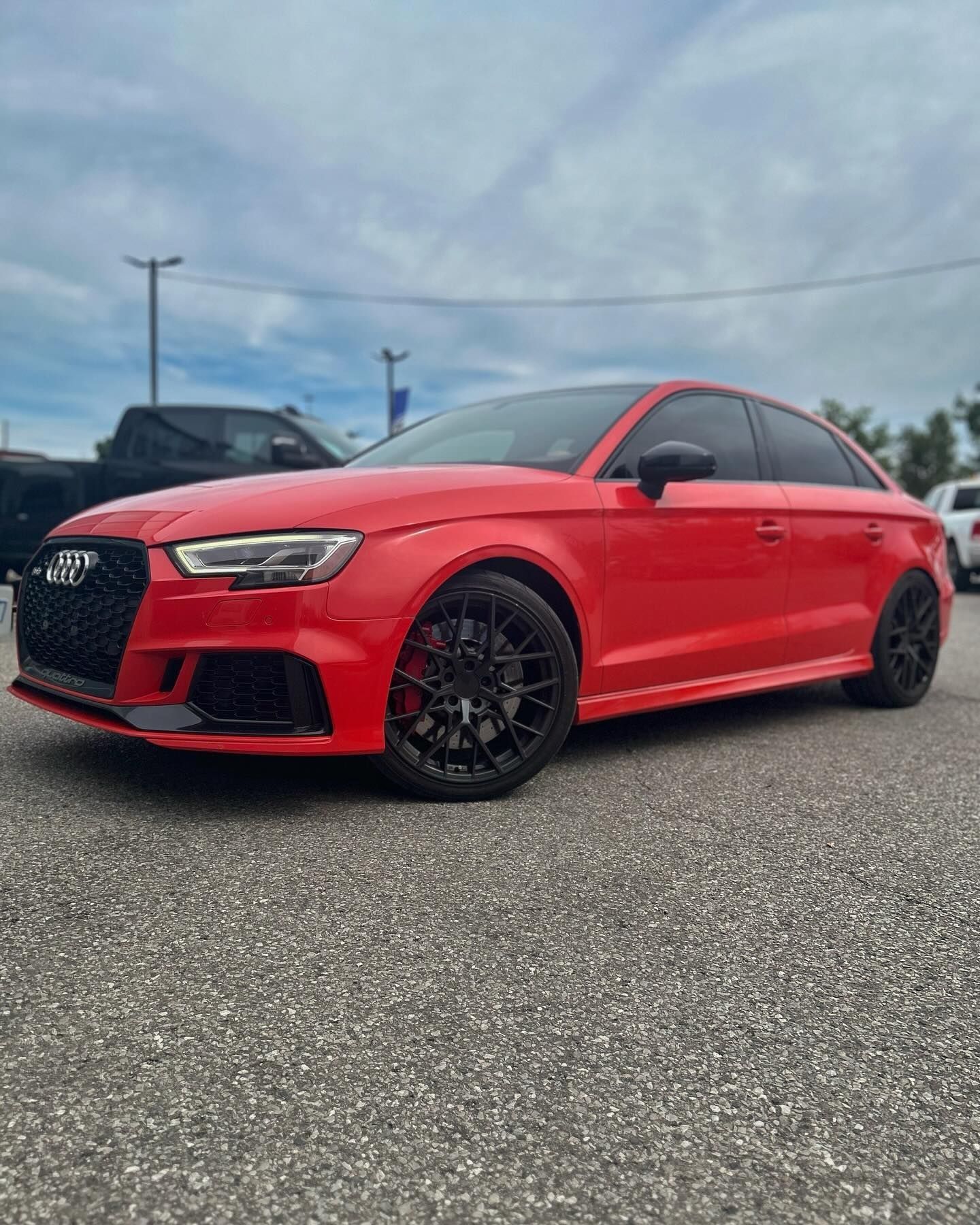 A bright red Audi sedan parked in a paved lot under a cloudy sky, featuring black wheels and a black front grille.