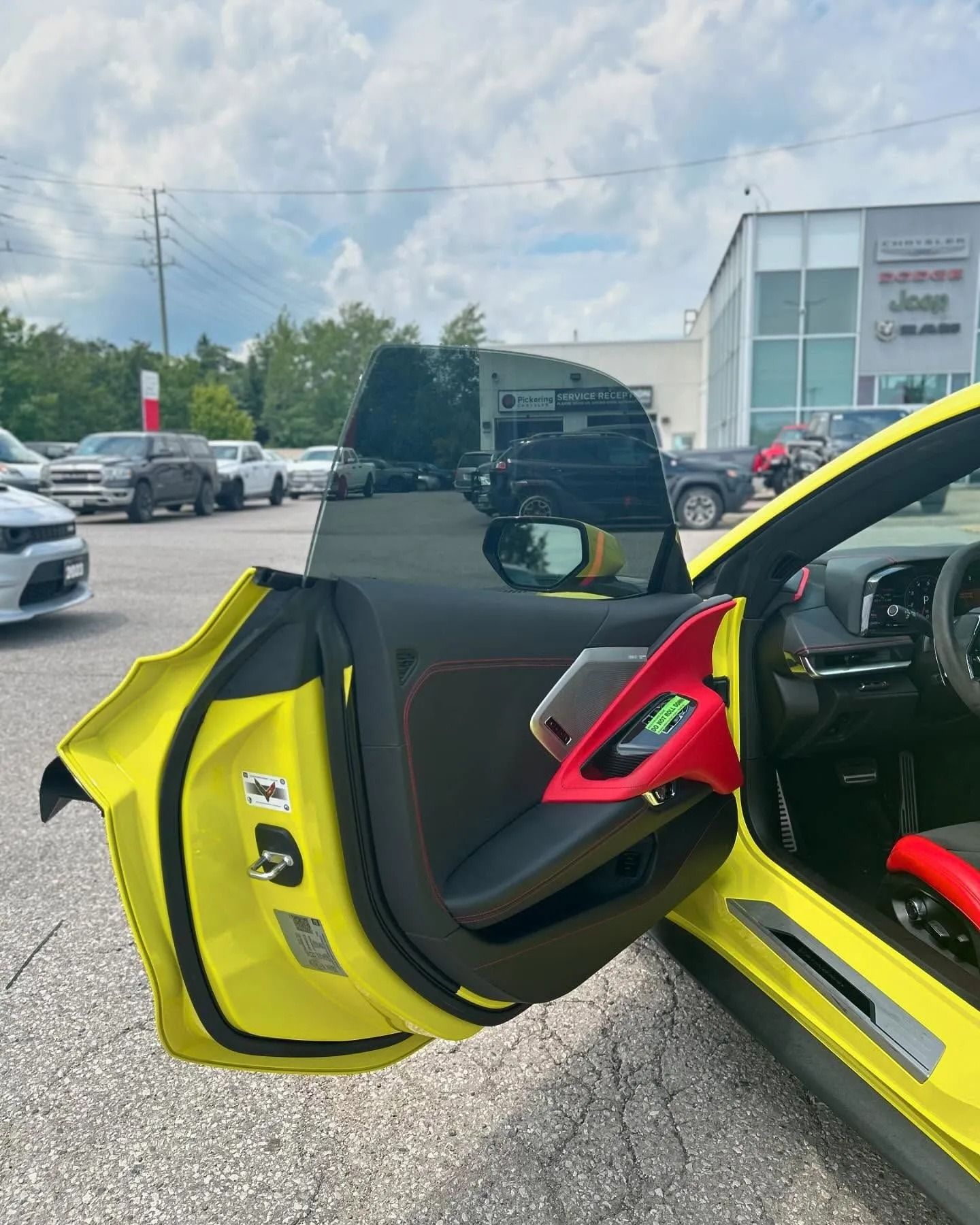 The open driver-side door of a yellow Chevrolet Corvette reveals a black and red interior at a car dealership.