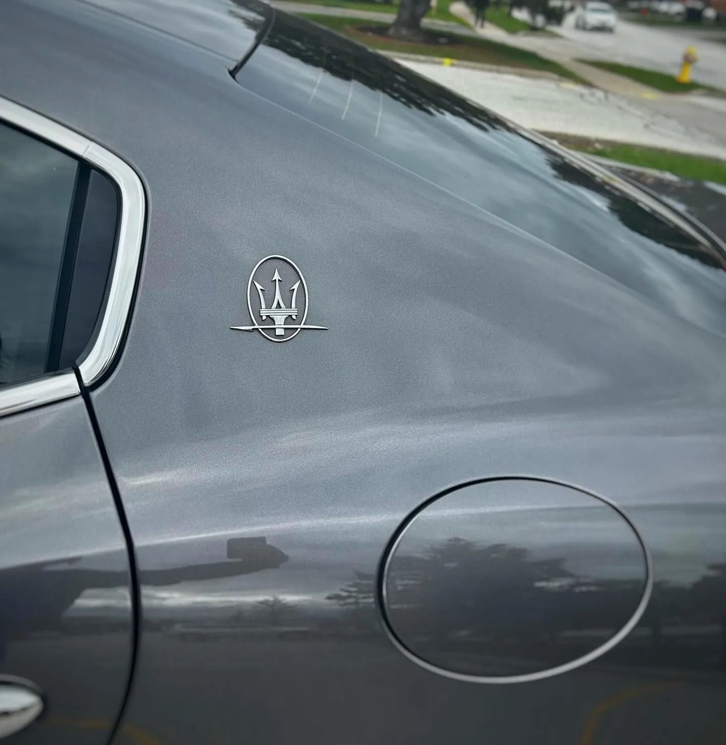 A close-up of a silver Maserati trident emblem on the rear pillar of a dark gray car, positioned above the fuel door.