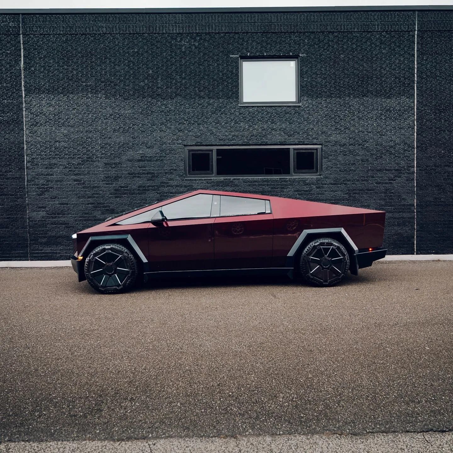 A dark red Tesla Cybertruck parked on a gravel lot in front of a dark, textured building.