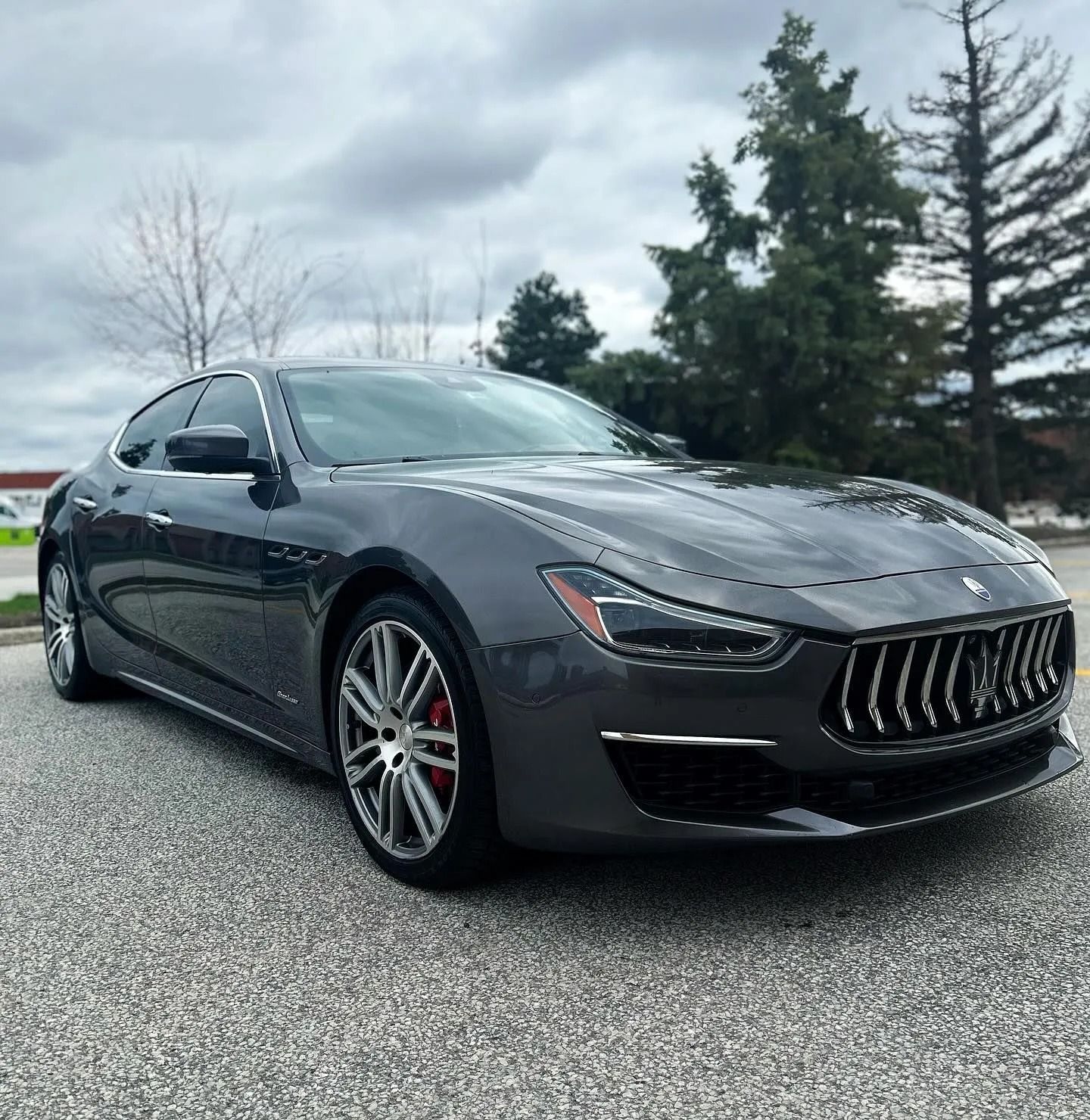 A dark grey Maserati Ghibli sedan parked on a gravel lot under a cloudy sky.