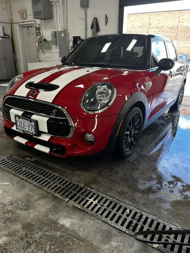 A red Mini Cooper with white racing stripes parked inside a bright garage with a floor drain.