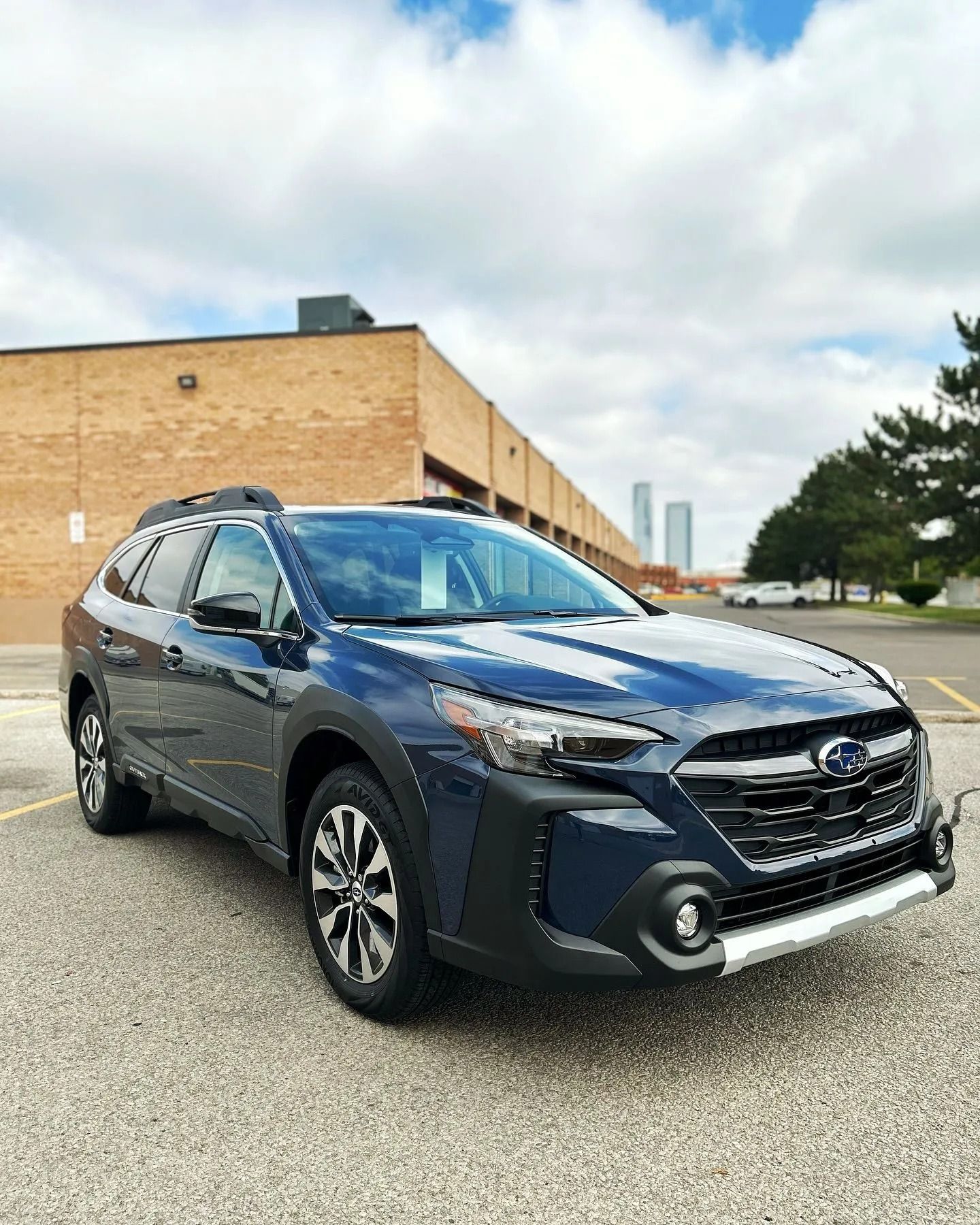 A dark blue Subaru Outback parked in a paved lot in front of a tan brick building under a cloudy sky.