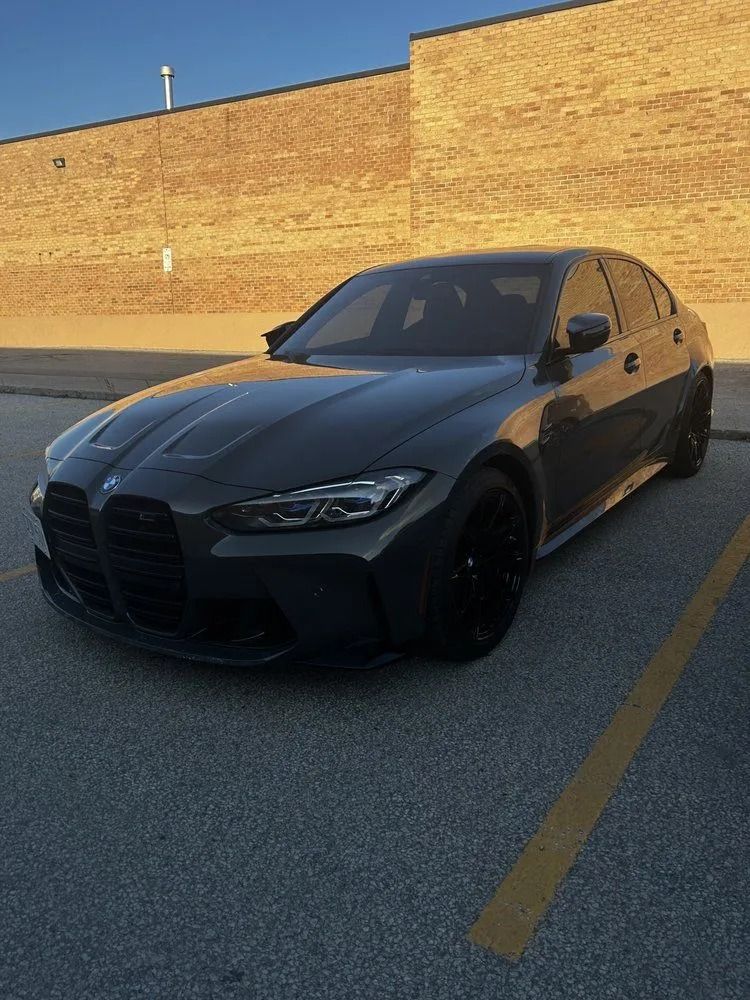 A dark gray BMW M3 sedan parked in a parking lot next to a brick wall.