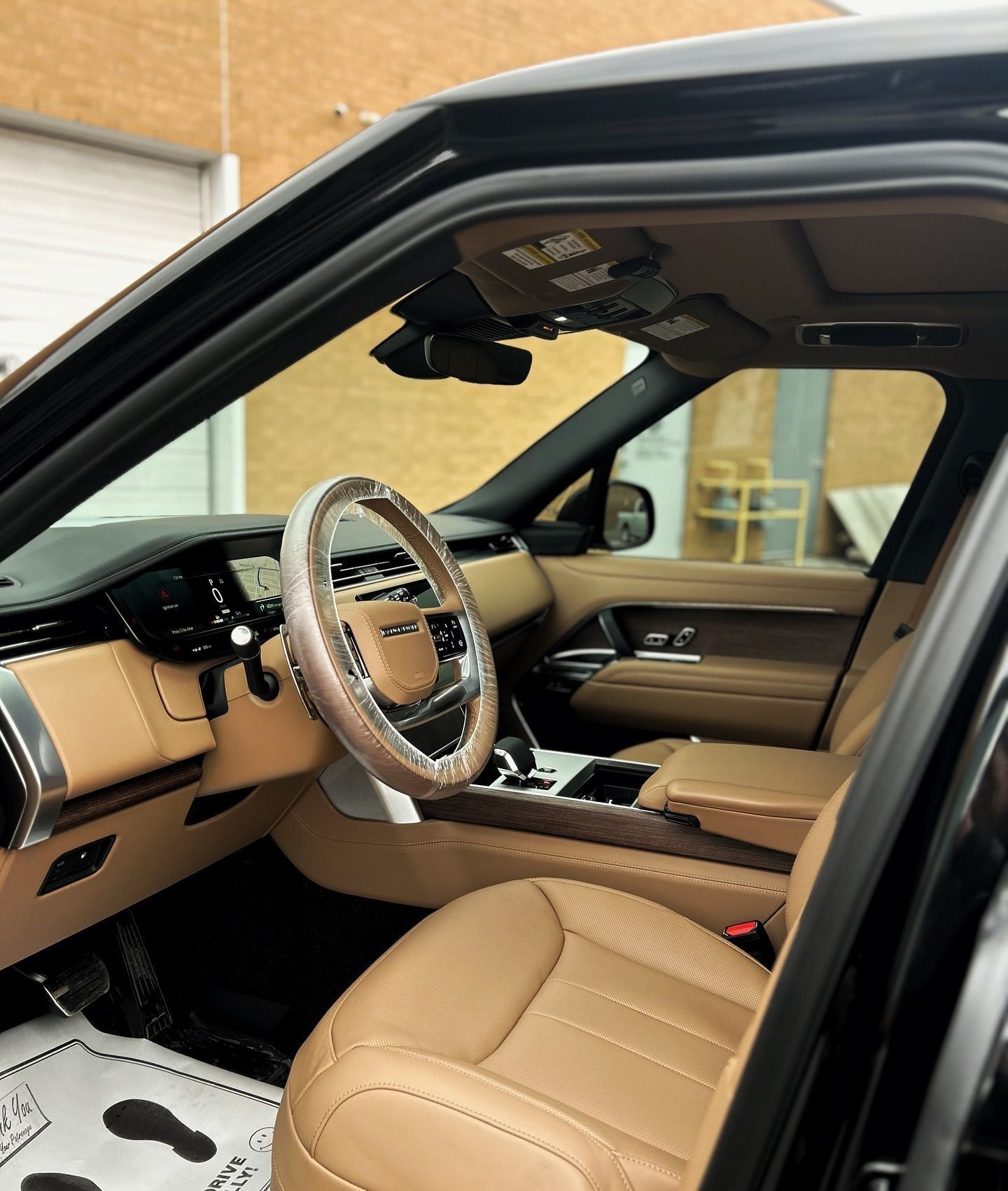 Interior view of a vehicle with tan leather seats and dashboard, featuring a steering wheel covered in protective film.