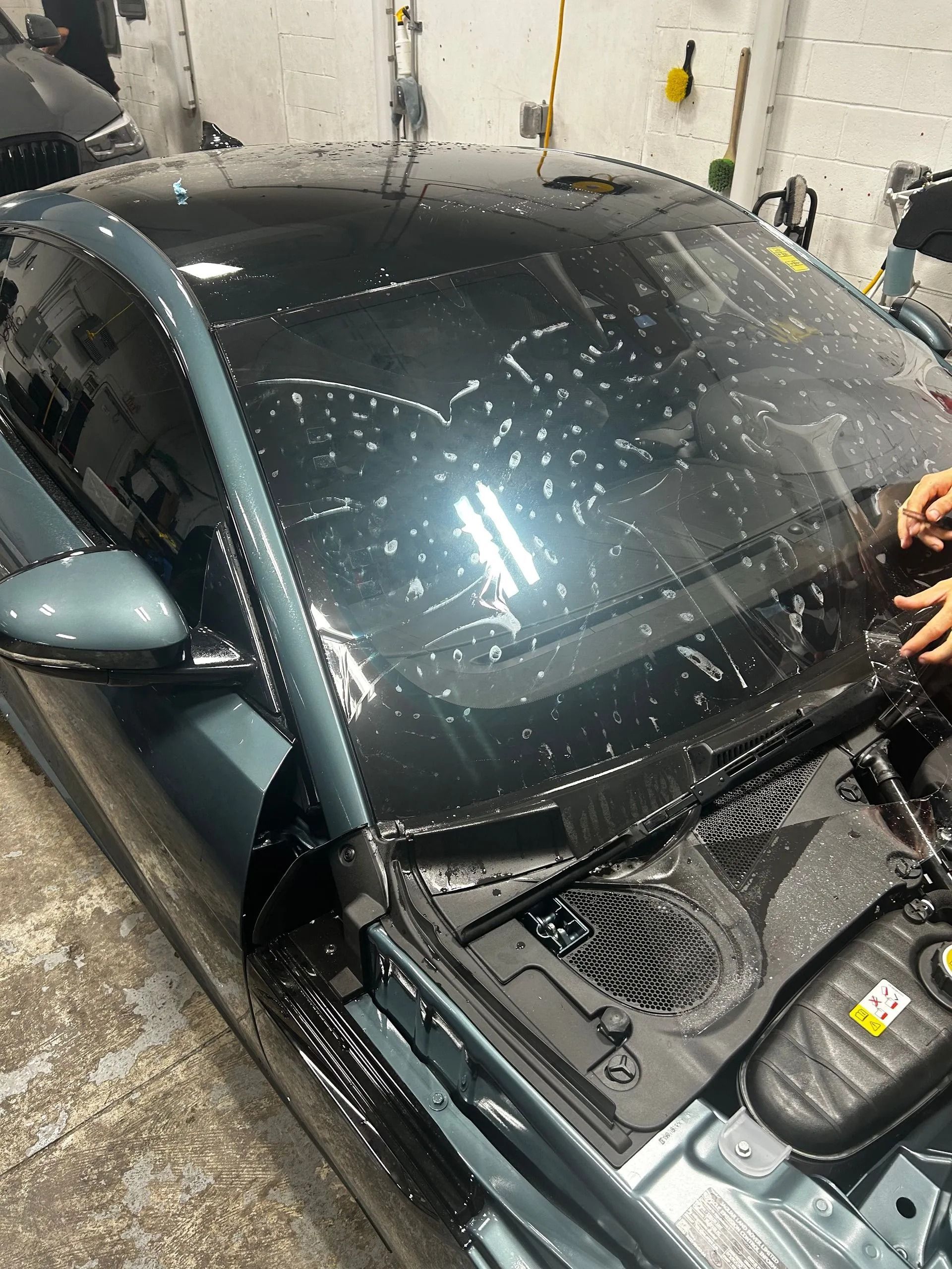 A person installing protective film on the dark-tinted windshield of a gray car inside a garage.