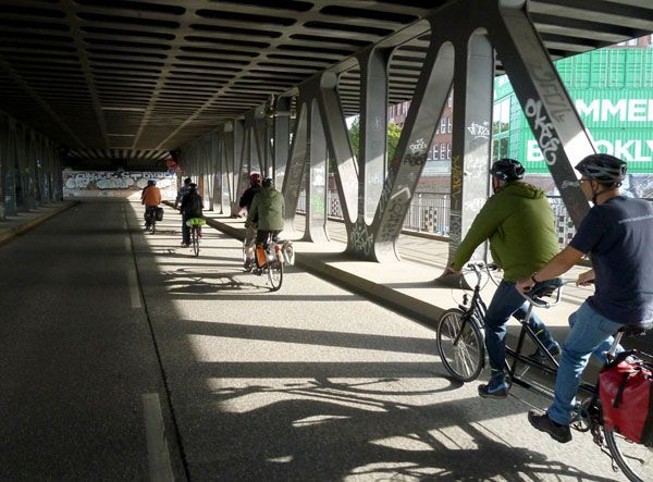 Das Foto zeigt Tandemfahrer vom Verein, die auf der Eisenbrücke über einen Hafenarm fahren nah an den Deichtorhallen. Die Tandemfahrer fahren auf der unteren Ebene, darüber ist ein Metalldach, weil dort die Bahnstrecke Hauptbahnhof-Harburg läuft.