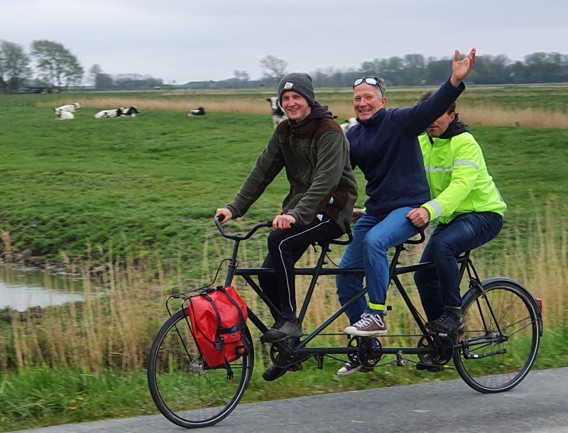 Das Foto zeigt die drei Männer auf dem Tandem. Im Hintergrund Weidegebiete mit Kühen. Am Gepäckträger vorne hängt eine rote Fahrradtasche mit Getränken und Keksen.