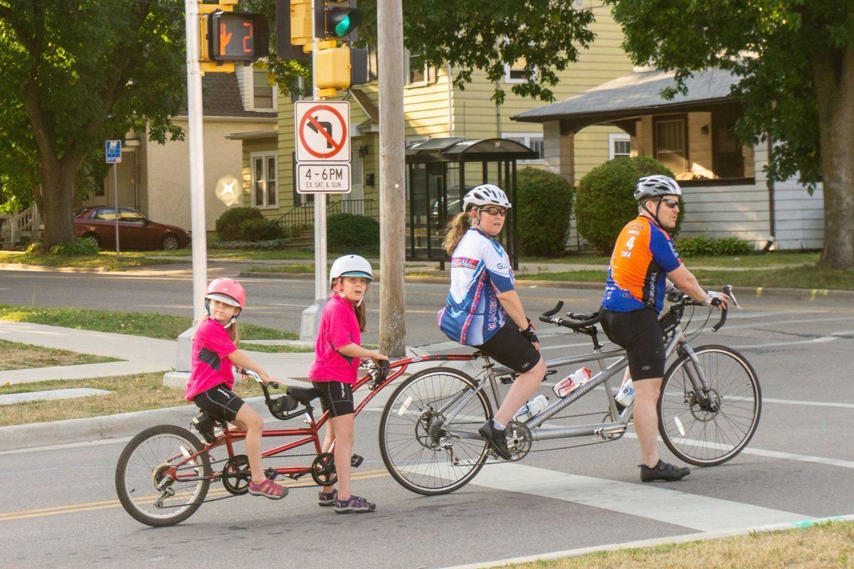 Das Bild zeigt ein Renntandem. An der Sattelstütze hinten is ein Fahrradanhänger gekoppelt. Der Anhänger erweitert das Tandem um zwei weiteren Sitzplätze inklusive Predalen. Es handelt sich offensichtlich um eine Familie: der Vater vorne, dann die Mutter und auf dem Anhängererweiterung sitzen zwei Mädchen von geschätzt 7 oder 9 Jahre alt. Alle sind in Rennradkleidung. Der Hintergrund lässt vermuten, dass das Bild in den Vereinigten Staten aufgenommen wurde.