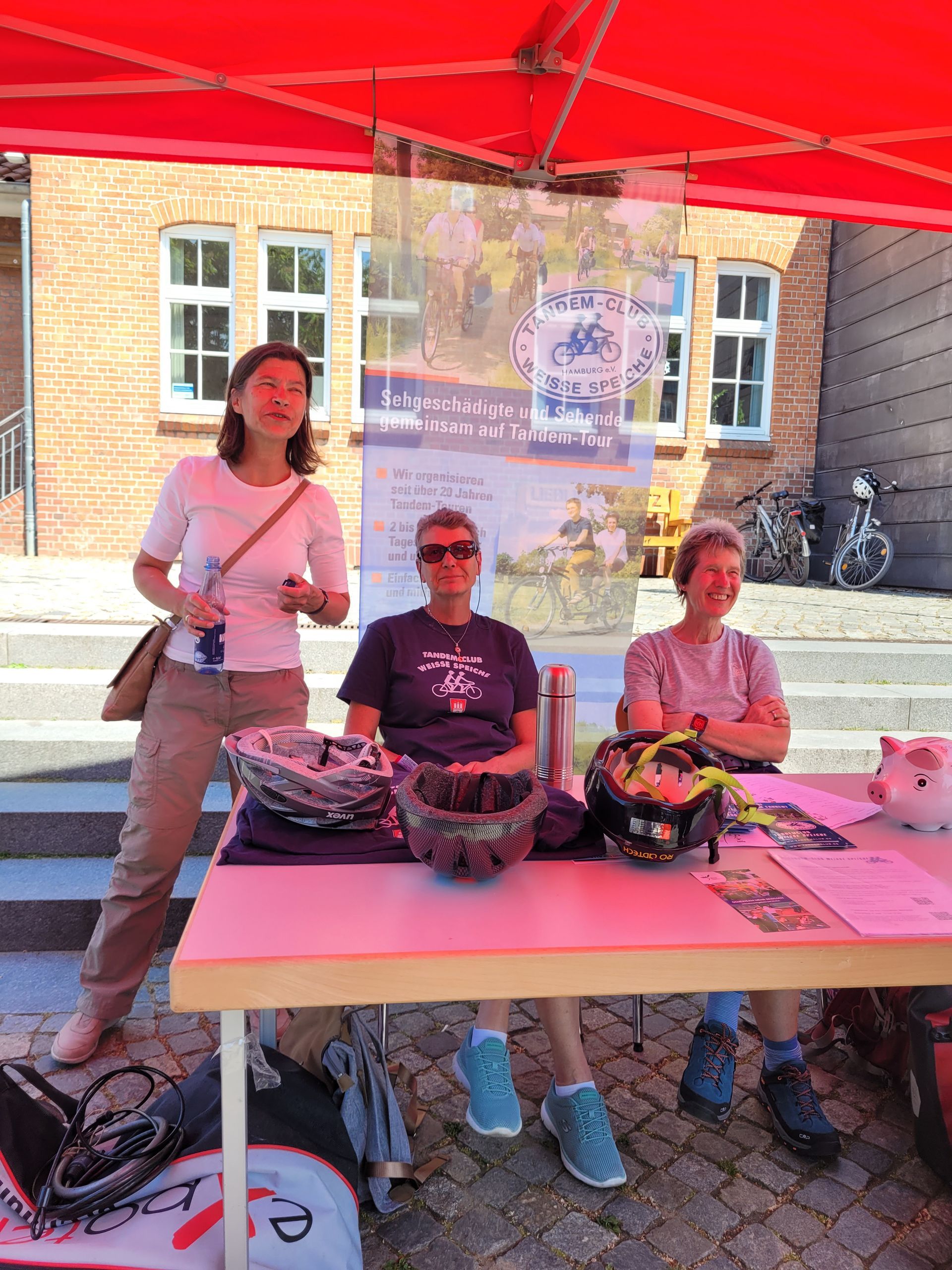 Das Bild zeigt ein Foto von unserem Marktkram auf dem Markt. Drei Tandemfahrerinnen unseres Clubs lachen die Besucher freundlich zu.