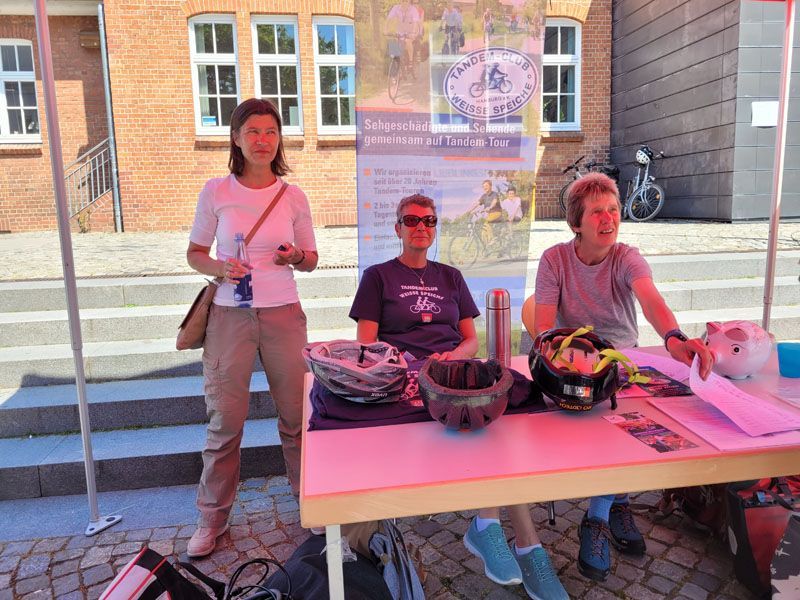 Das Foto zeigt ein den Marktkram vom Tandemclub. Da die Sonne scheint und die Plastic Zeltdecke rot ist, scheint der ganzen Marktkram etwas rötlich. Drei Vereinsmitglieder stehen und sitzen hinter dem Tisch mit Infomaterial.