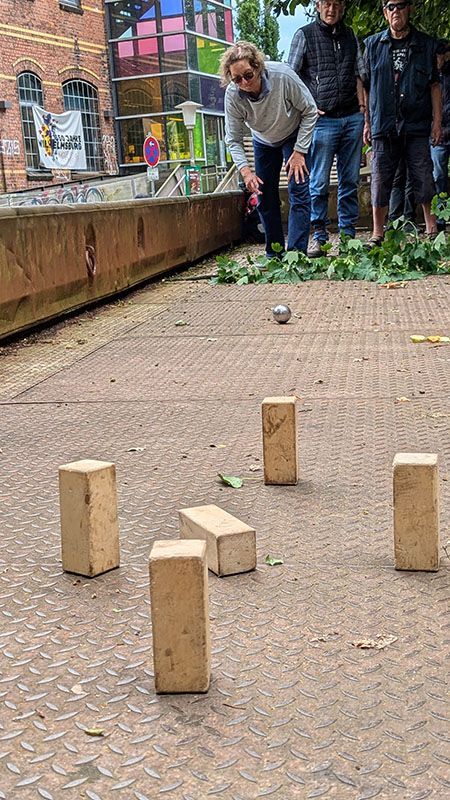 Flacher, offener Güterwagen mit ganz niedrigen Wänden. Darauf stehen hinten im Bild einige Tandemfahrer hinter einen Zweig, der als Linie verwendet wird. Gabi wirft gerade einen Boule Spiel Kugel. Vorne im Bild die Holzklötze, die umgeworfen werden müssen. Ein Holzklotz ist schon umgefallen vom vorherigen Wurf.