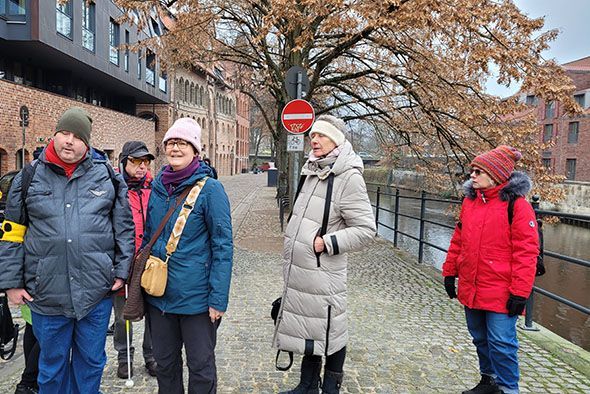 Foto von Lüneburg mit einem Teil der Wandergruppe darauf. Sie stehen auf dem Kopfsteinpflaster an einem alten Kanal. Darum herum alte Häuser aus Backsteinen.