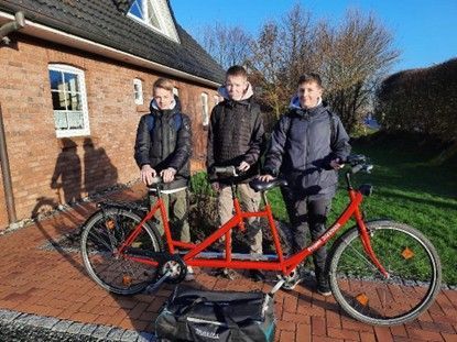 Drei 9. Klassler mit warmen Jacken an stehen stolz hinter ihrem roten Tandem im Garten eines Hauses in Viöl.