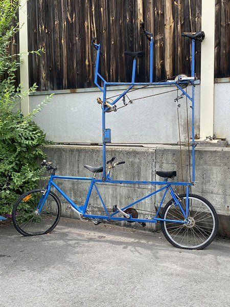 Das Foto zeigt ein etwas besonderes Tandem mit Platz für vier Personen. Im Gegensatz zu einem normalen Tandem sitzen hier unten zwei Personen und oben auch zwei. Alle Pedale sind mittels Ketten verbunden. Das Fahrrad ist gegen ein Gebäude gelehnt und es ist unbekannt, ob man darauf fahren kann.