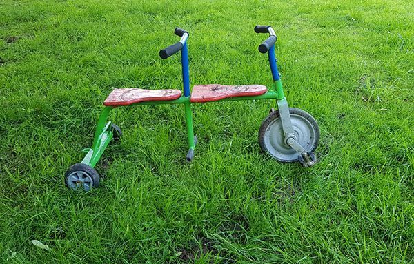 Das fotografierte Dreiradtandem, das im Text beschrieben wird. Das Vorderrad hat direkt montierte Pedale, wie beim Kinderdreirad. Unter dem zweiten Lenker gibt es nur eine Stange, auf dem die Füße ruhen können. Das Dreiradtandem steht auf einer grünen Wiese.