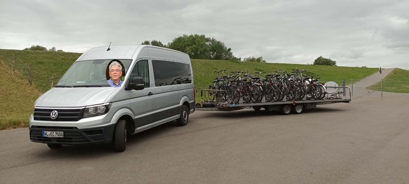 Das Foto zeigt eine Landschaft mit einem Deich. Vor dem Deich ist ein Personenbus mit Tandemanhänger abgebildet. Dort, wo man durch die Frontscheibe des Buses den Fahrer sehen könnte, ist ein Profilfoto (Frontansicht) von Hans aufgeklebt.
