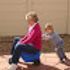 Woman in pink top sits with a toddler, who leans on her back, outdoors.